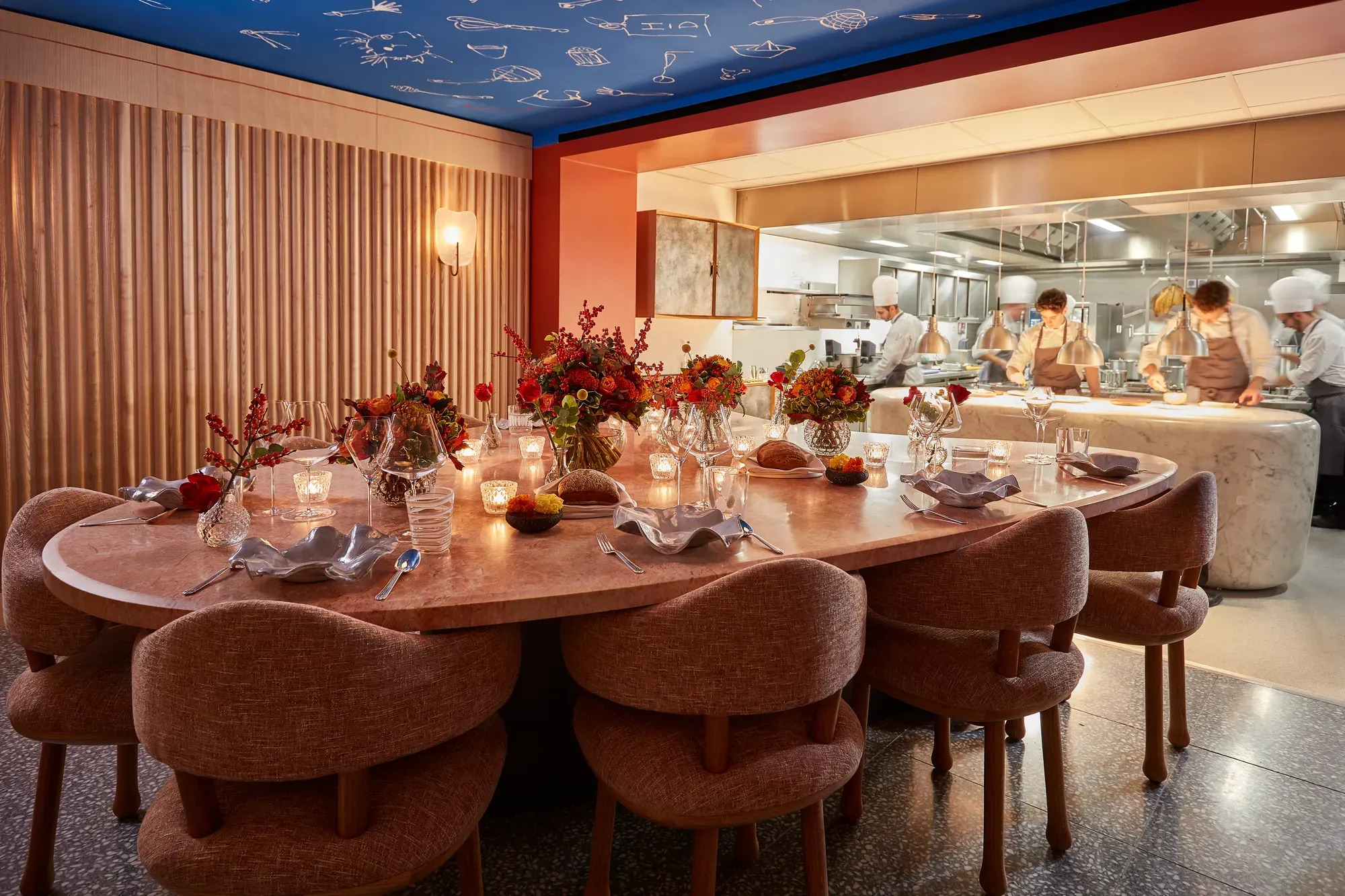 Candlelit chef’s table set with flowers beside an open kitchen where chefs prepare dishes.