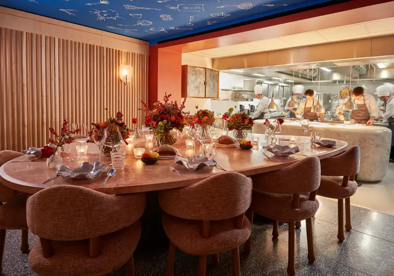 Candlelit chef’s table set with flowers beside an open kitchen where chefs prepare dishes.