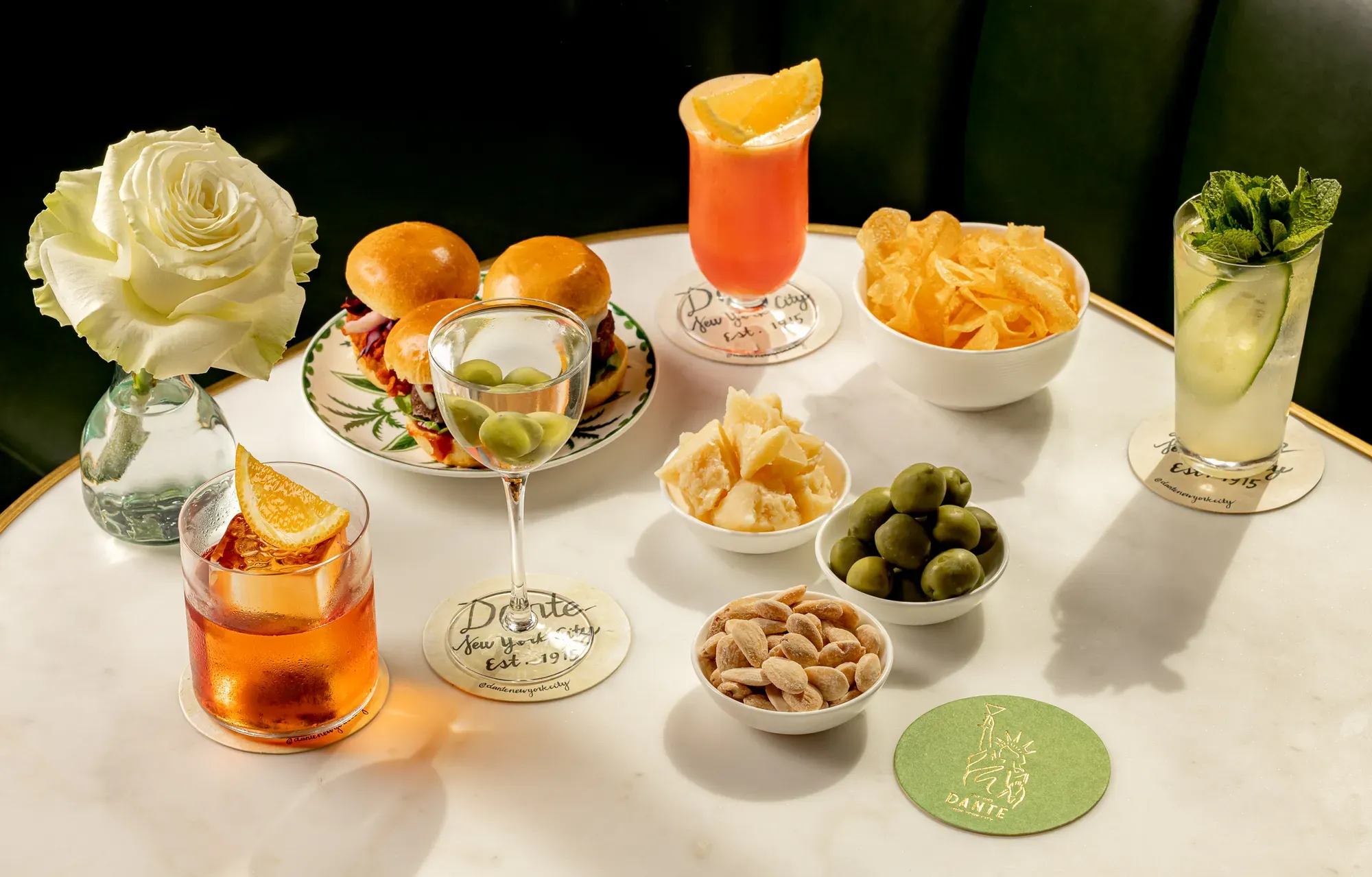 Cocktails, sliders, and bar snacks arranged on a marble table with white rose centerpiece.