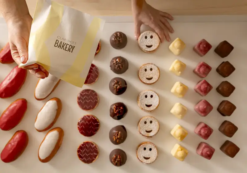 Hand holding a Claridge’s Bakery bag above a neatly arranged selection of colourful pastries.