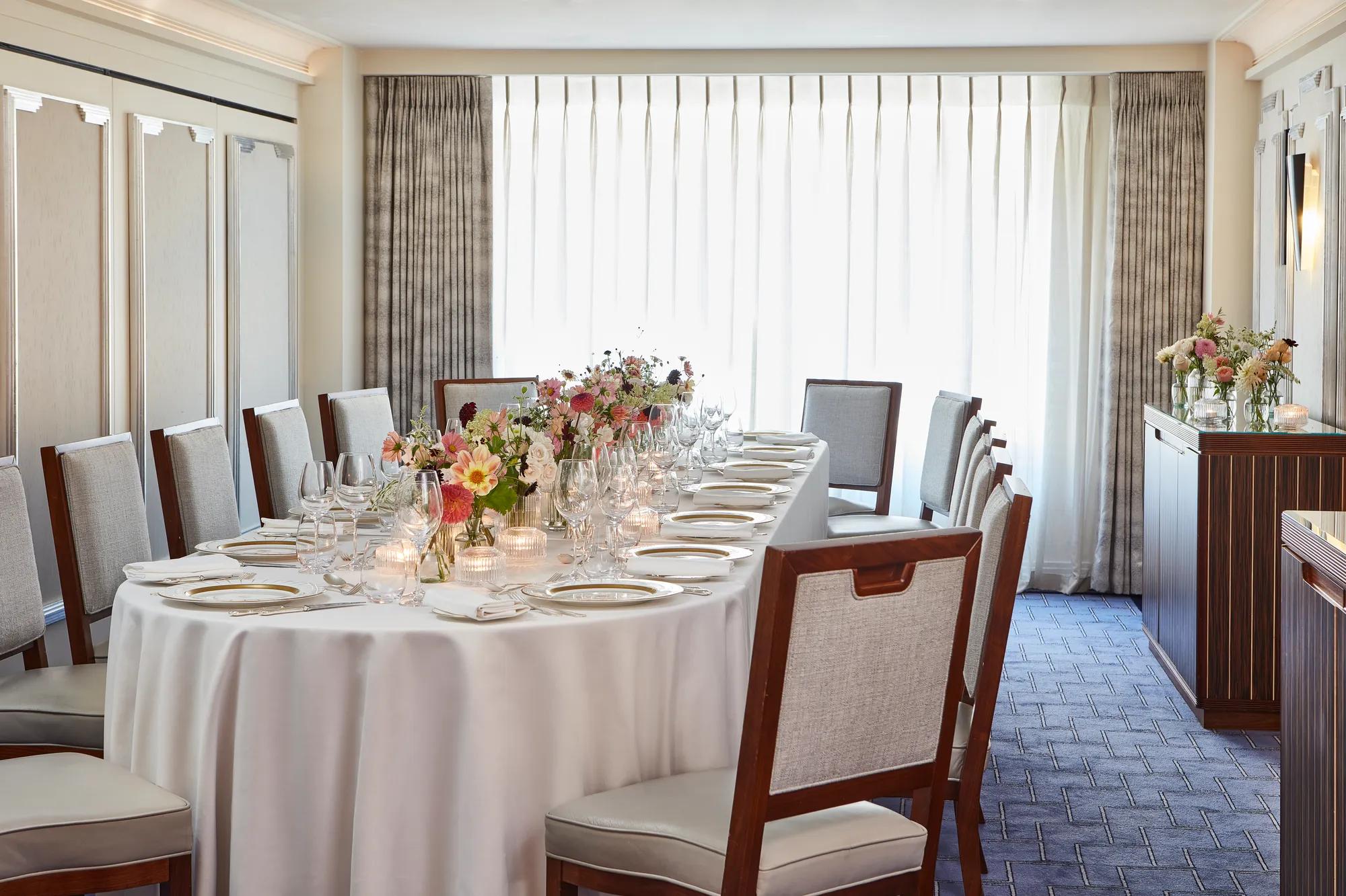 Long dining table set with white linens, glassware, candles, and floral centrepieces in a bright private room with curtains