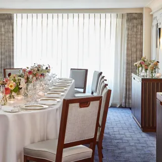 Long dining table set with white linens, glassware, candles, and floral centrepieces in a bright private room with curtains
