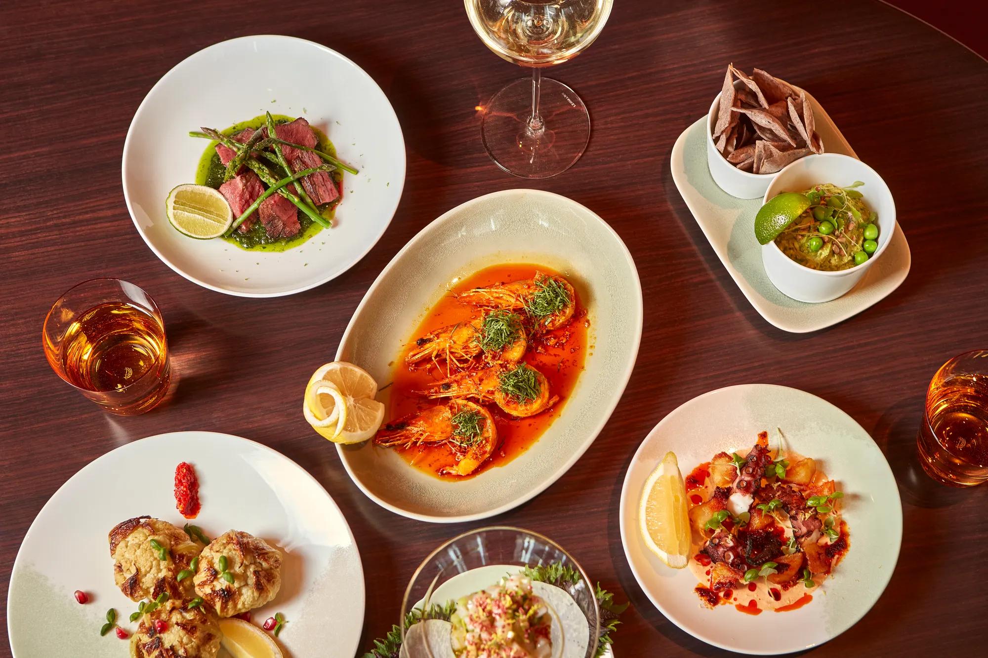Overhead view of assorted seafood dishes on a wooden table, including prawns, tuna, ceviche, small plates, and drinks.
