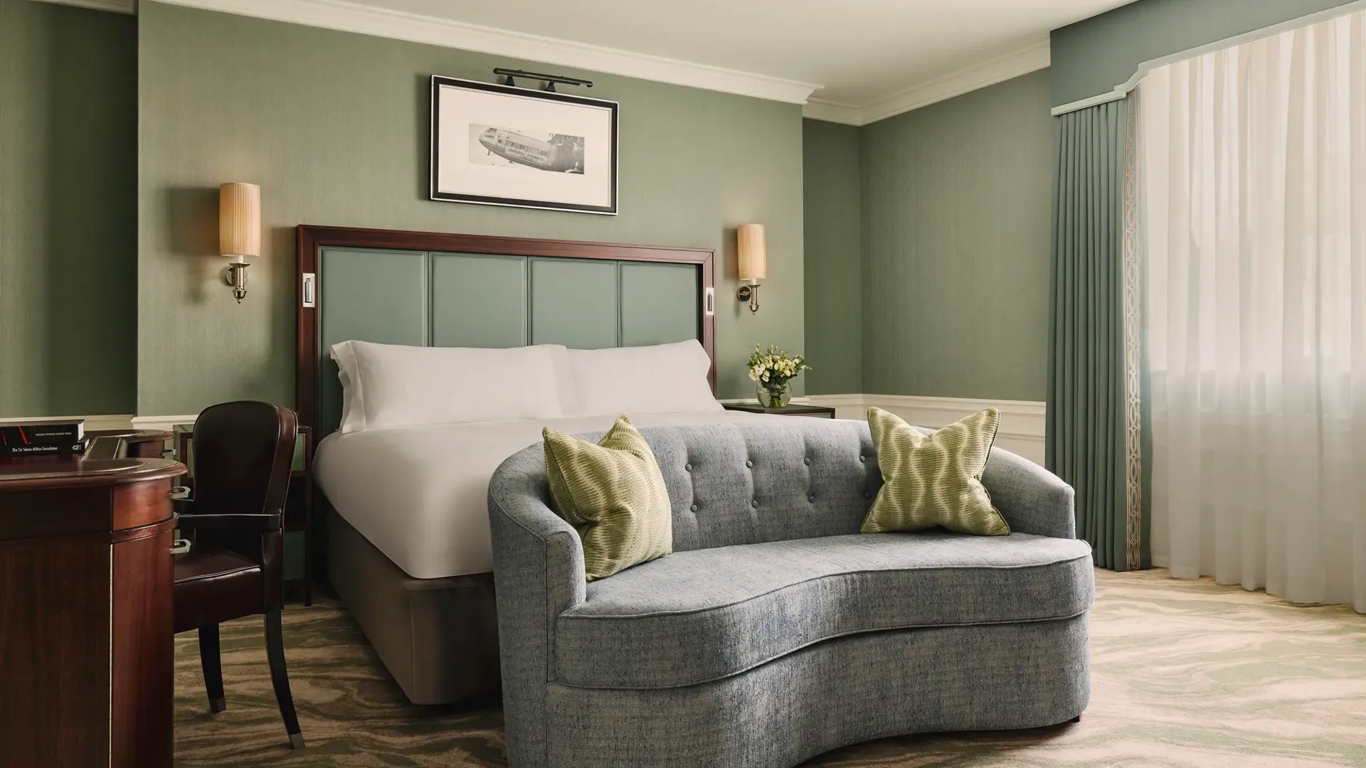 Calm bedroom with a king bed, soft green walls, a curved blue loveseat at the foot of the bed, and sheer curtains letting in daylight.