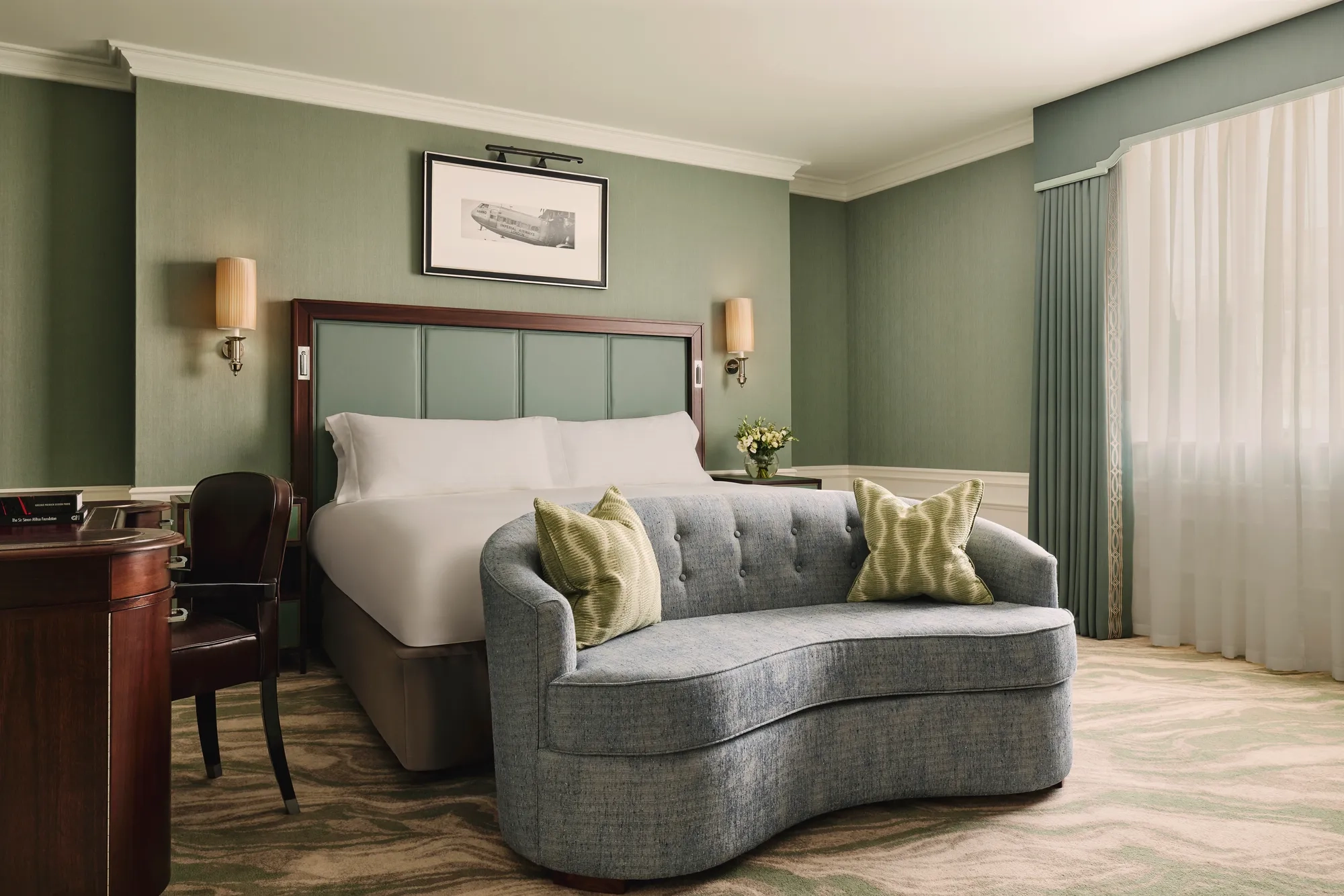Calm bedroom with a king bed, soft green walls, a curved blue loveseat at the foot of the bed, and sheer curtains letting in daylight.
