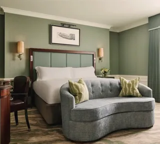 Calm bedroom with a king bed, soft green walls, a curved blue loveseat at the foot of the bed, and sheer curtains letting in daylight.