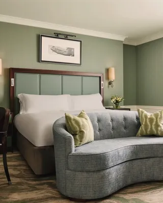 Calm bedroom with a king bed, soft green walls, a curved blue loveseat at the foot of the bed, and sheer curtains letting in daylight.