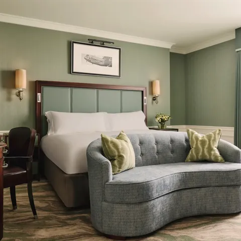 Calm bedroom with a king bed, soft green walls, a curved blue loveseat at the foot of the bed, and sheer curtains letting in daylight.