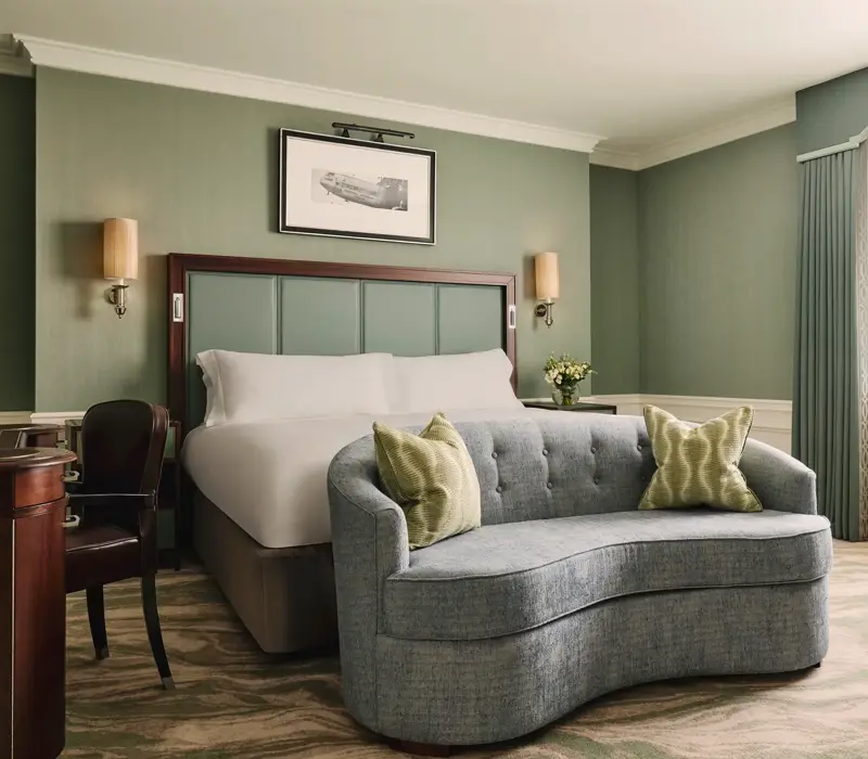 Calm bedroom with a king bed, soft green walls, a curved blue loveseat at the foot of the bed, and sheer curtains letting in daylight.