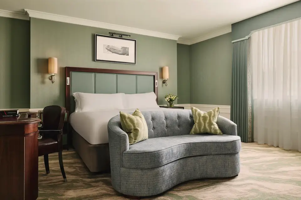 Calm bedroom with a king bed, soft green walls, a curved blue loveseat at the foot of the bed, and sheer curtains letting in daylight.
