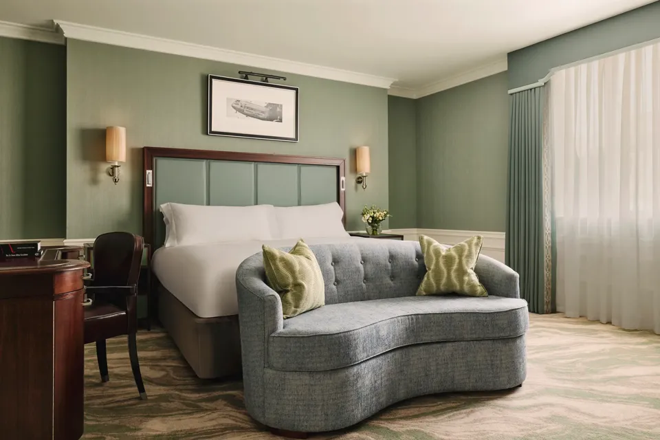 Calm bedroom with a king bed, soft green walls, a curved blue loveseat at the foot of the bed, and sheer curtains letting in daylight.