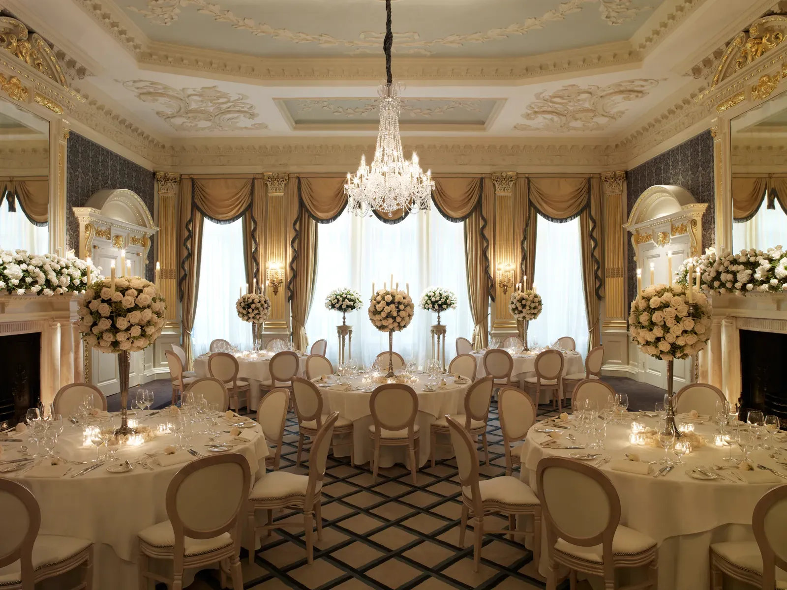 An ornate ballroom set for a formal dinner, featuring round tables with white linens, tall floral centrepieces of white roses, and a grand crystal chandelier beneath an intricately detailed ceiling with gold accents and large draped windows.