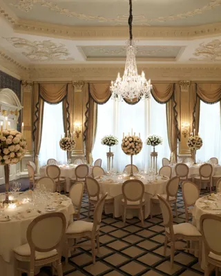 An ornate ballroom set for a formal dinner, featuring round tables with white linens, tall floral centrepieces of white roses, and a grand crystal chandelier beneath an intricately detailed ceiling with gold accents and large draped windows.