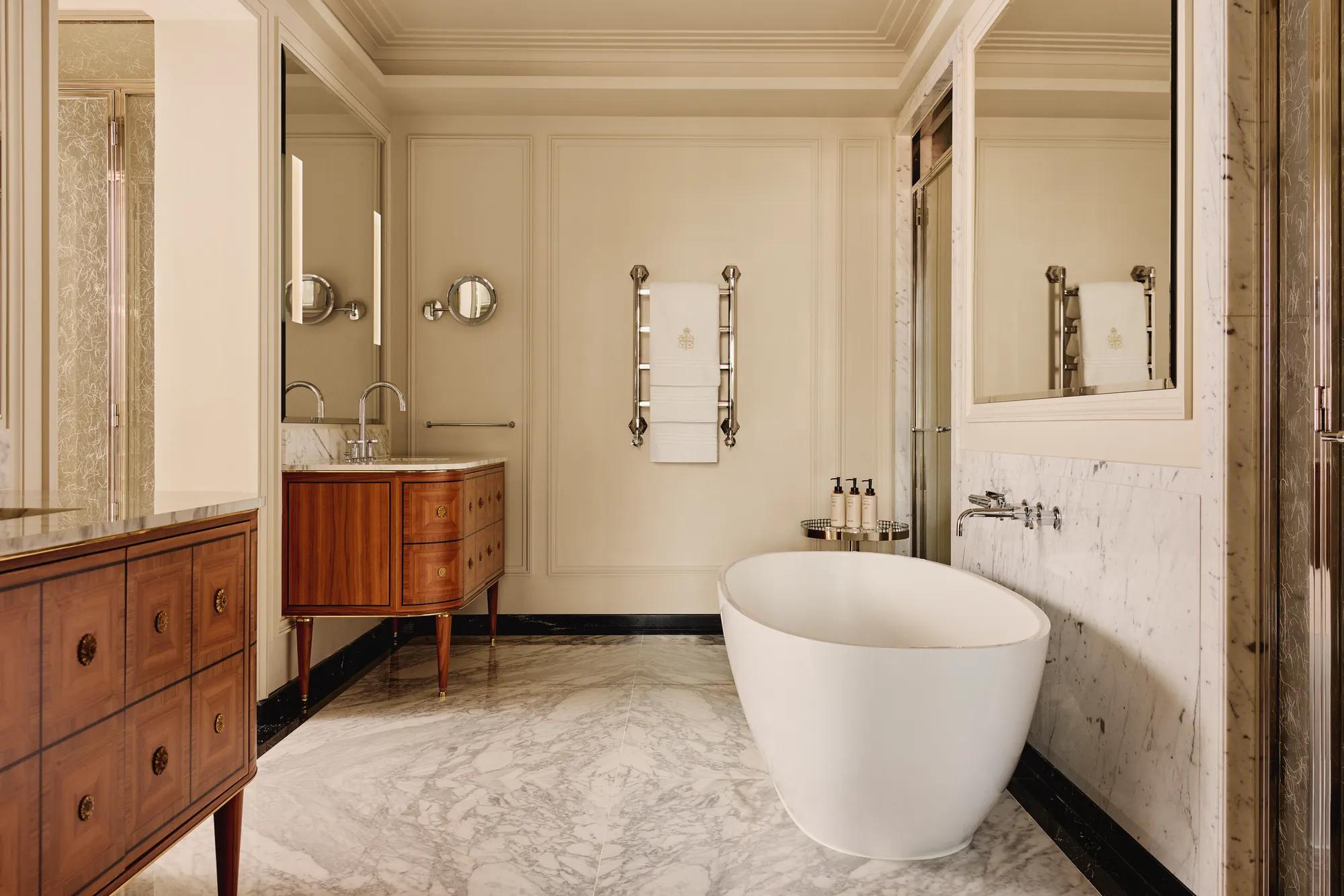 Refined marble bathroom with a freestanding white bathtub, twin wooden vanities with marble tops, and chrome fixtures, featuring a heated towel rail and large wall mirrors.