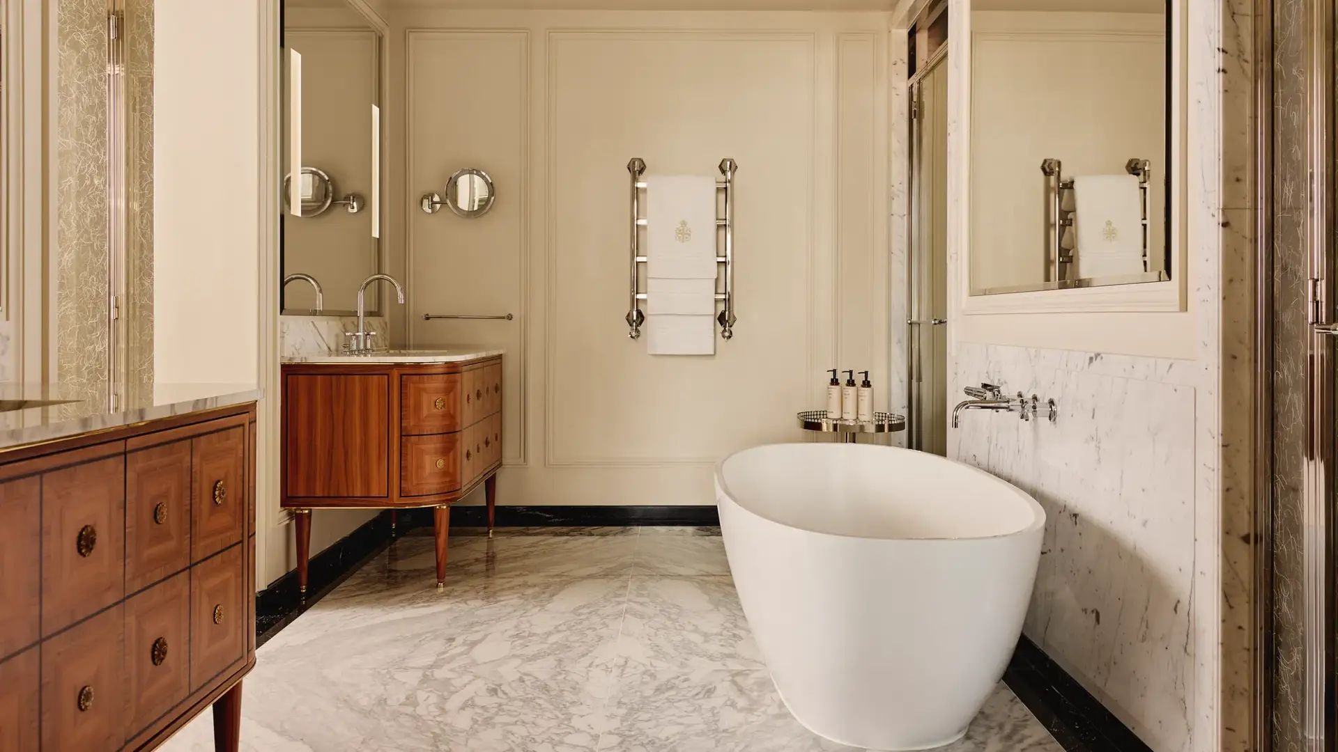 Refined marble bathroom with a freestanding white bathtub, twin wooden vanities with marble tops, and chrome fixtures, featuring a heated towel rail and large wall mirrors.