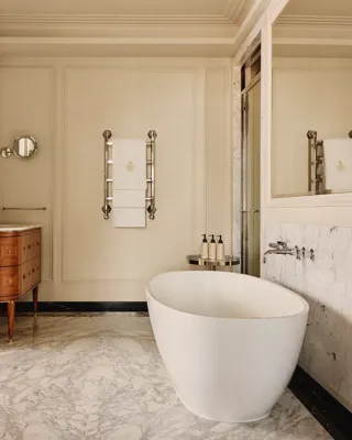 Refined marble bathroom with a freestanding white bathtub, twin wooden vanities with marble tops, and chrome fixtures, featuring a heated towel rail and large wall mirrors.