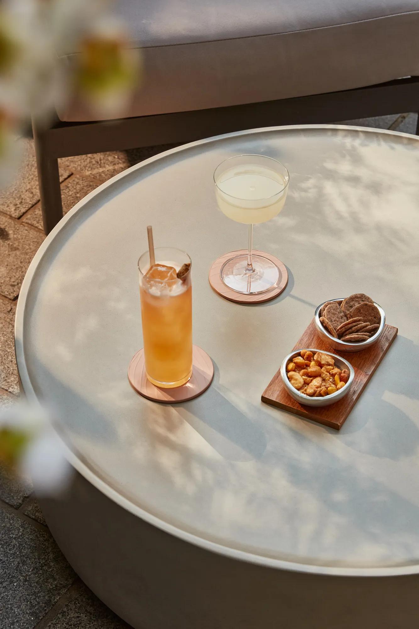 Outdoor table with a tall iced cocktail, coupe glass cocktail, mixed nuts, and crackers.