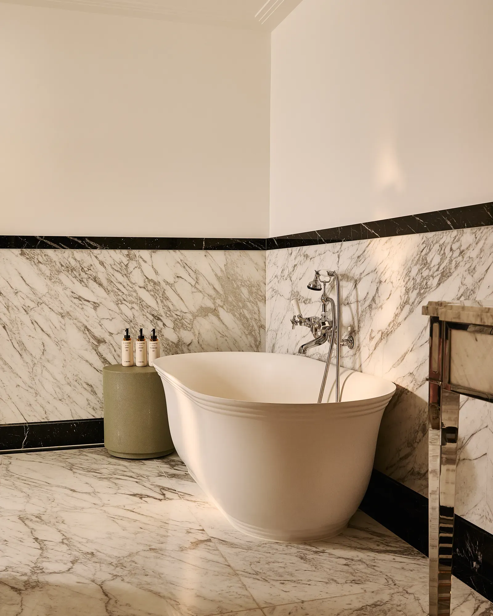 Marble bathroom with freestanding oval bathtub, chrome fixtures, stone side table with toiletries, and marble walls and floor.