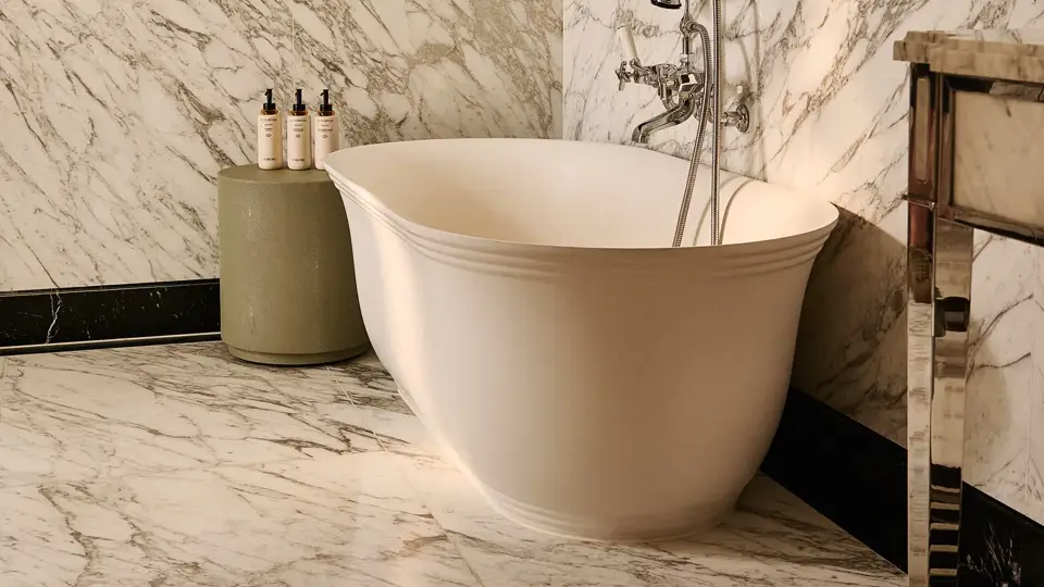 Marble bathroom with freestanding oval bathtub, chrome fixtures, stone side table with toiletries, and marble walls and floor.