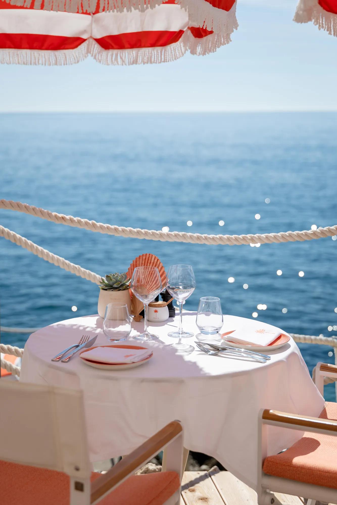Seaside dining table with white cloth, set for two beneath red-striped umbrellas overlooking the ocean.