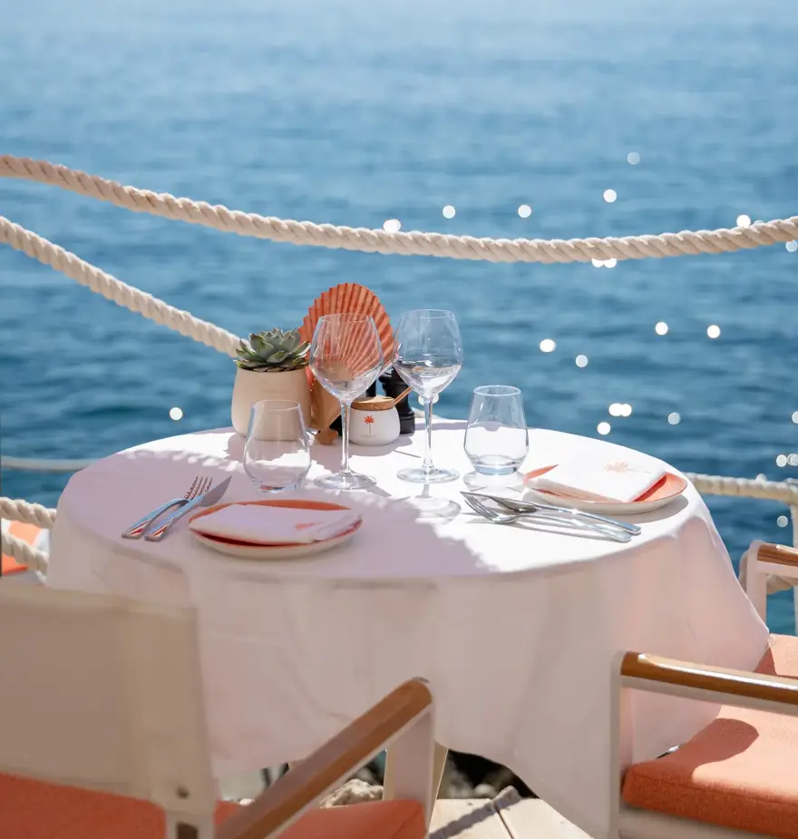 Seaside dining table with white cloth, set for two beneath red-striped umbrellas overlooking the ocean.