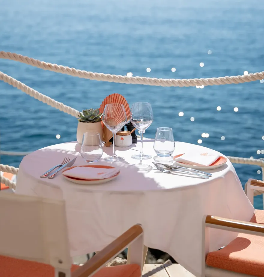 Table en bord de mer avec nappe blanche, dressée pour deux sous parasols rayés rouges face à l’océan
