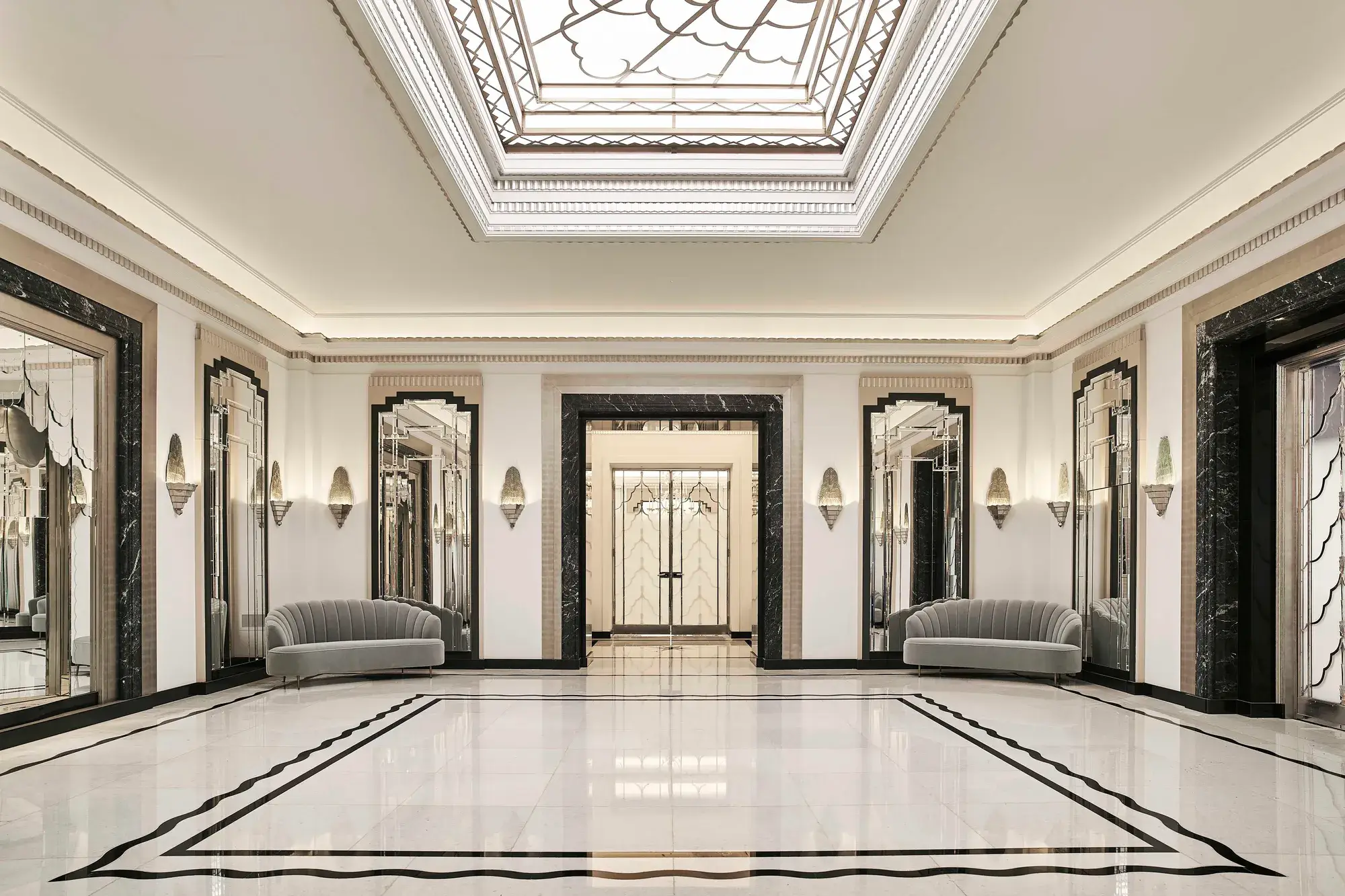 The Ballroom reception area at Claridge’s featuring polished marble floors, mirrored Art Deco wall panels, grey velvet sofas, and a large geometric skylight ceiling.