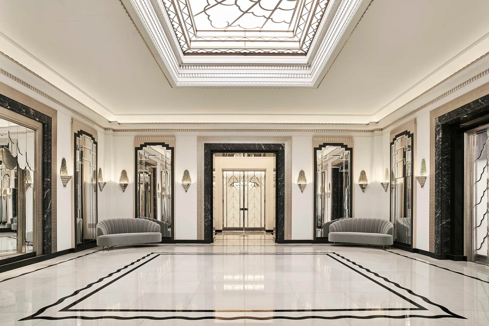 The Ballroom reception area at Claridge’s featuring polished marble floors, mirrored Art Deco wall panels, grey velvet sofas, and a large geometric skylight ceiling.