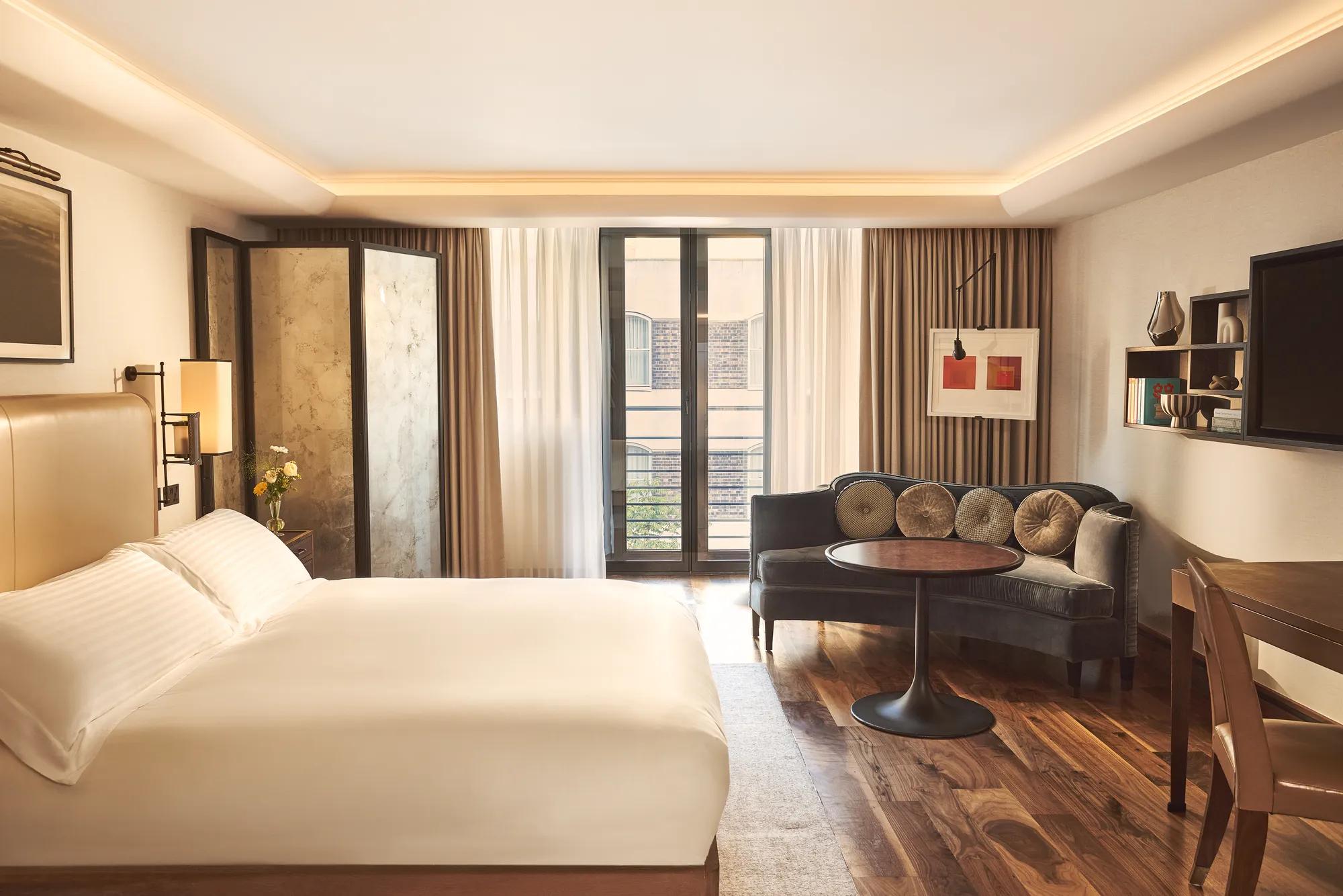 Bright bedroom with a cream bed, seating area with velvet sofa and round table, and floor-to-ceiling windows with sheer curtains.