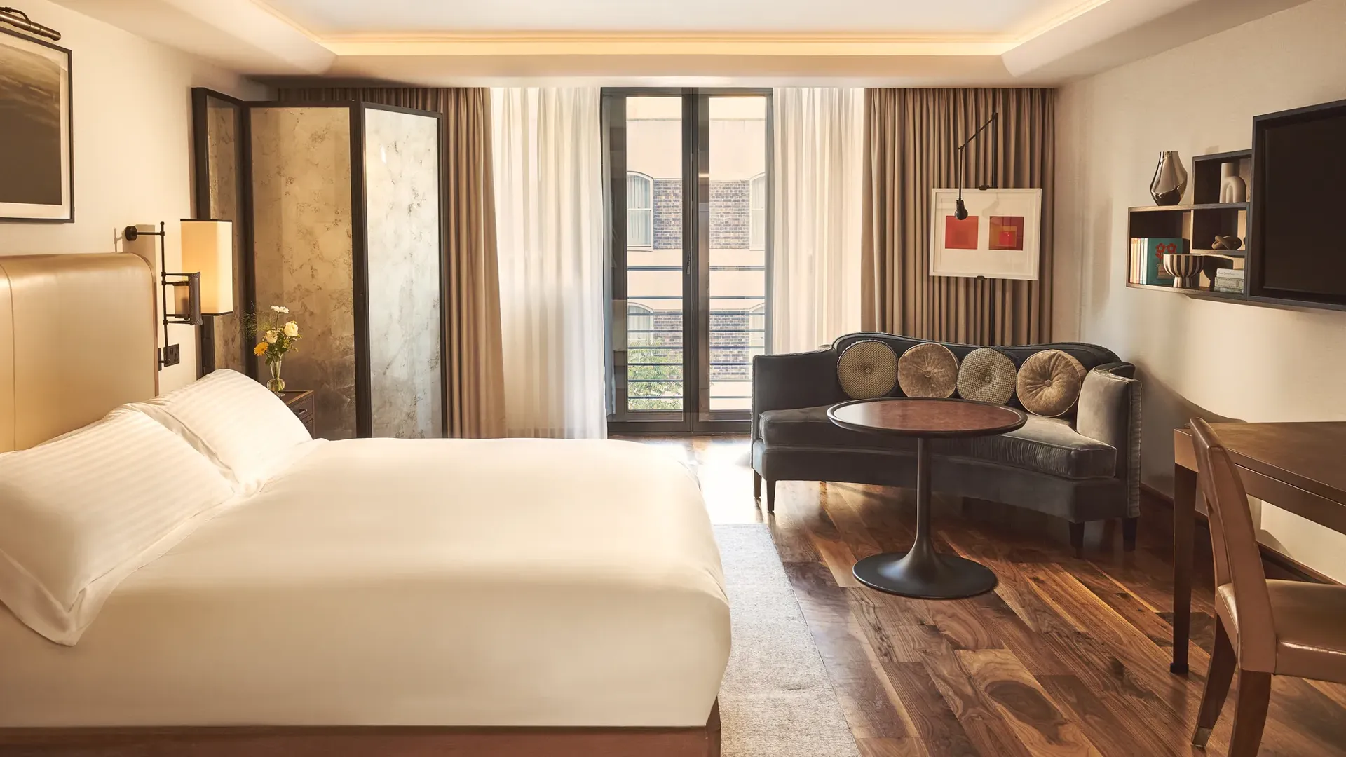 Bright bedroom with a cream bed, seating area with velvet sofa and round table, and floor-to-ceiling windows with sheer curtains.