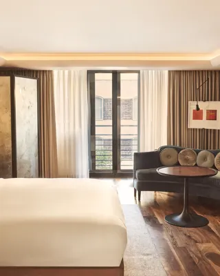 Bright bedroom with a cream bed, seating area with velvet sofa and round table, and floor-to-ceiling windows with sheer curtains.