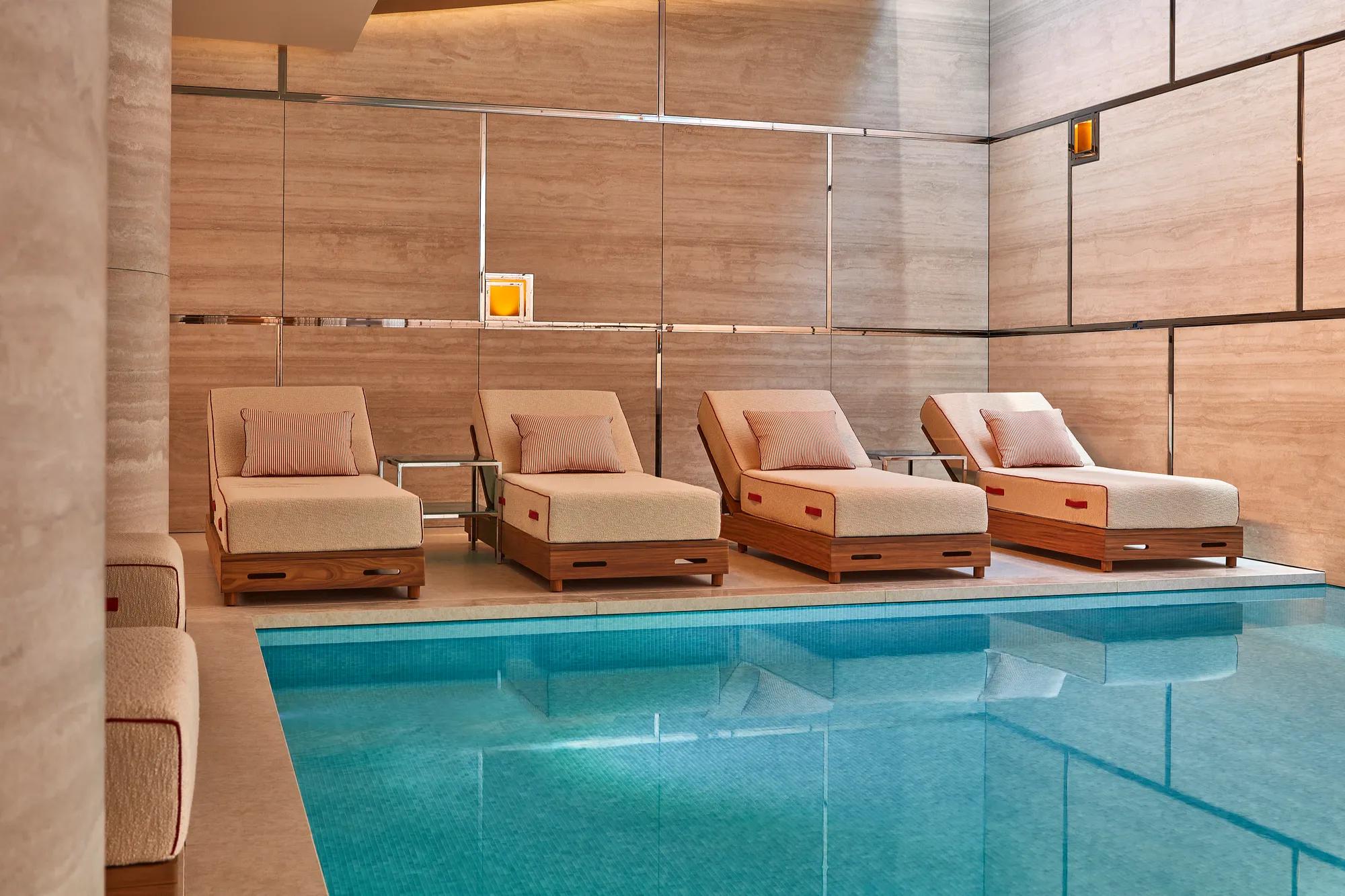 Four cushioned day beds with pillows beside an indoor pool, set against stone-panelled walls and soft lighting, creating a calm, spa-like relaxation area.