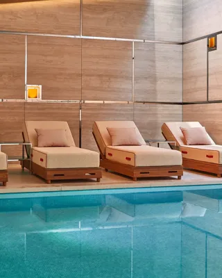 Four cushioned day beds with pillows beside an indoor pool, set against stone-panelled walls and soft lighting, creating a calm, spa-like relaxation area.
