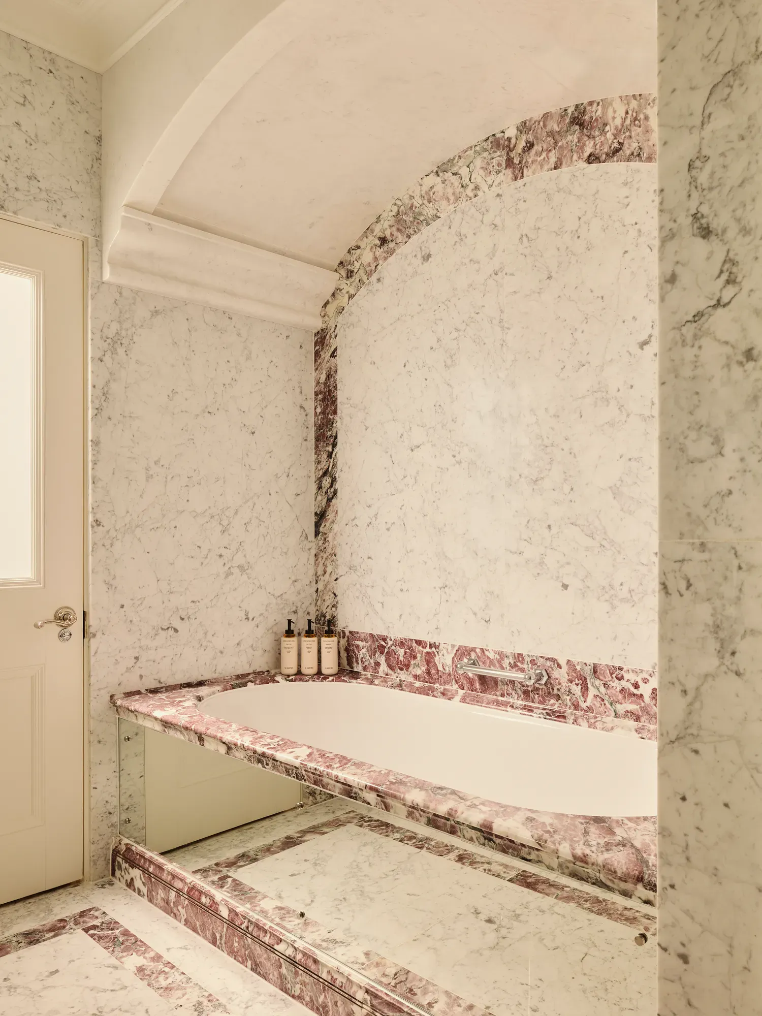 Marble bathroom with built-in tub, pink veining accents, and toiletries on the ledge.