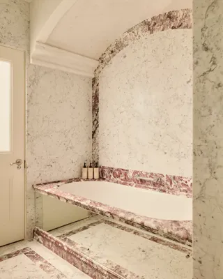 Marble bathroom with built-in tub, pink veining accents, and toiletries on the ledge.