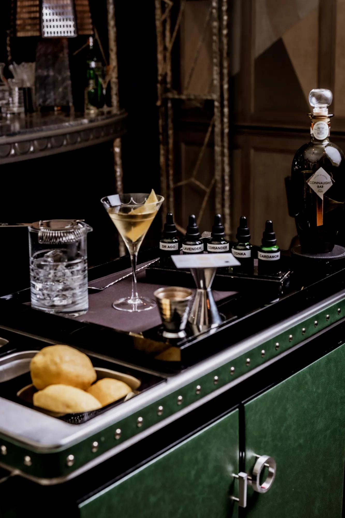 Martini trolley with mixing tools, citrus, bitters bottles, and a prepared martini beside a Connaught Bar gin bottle.