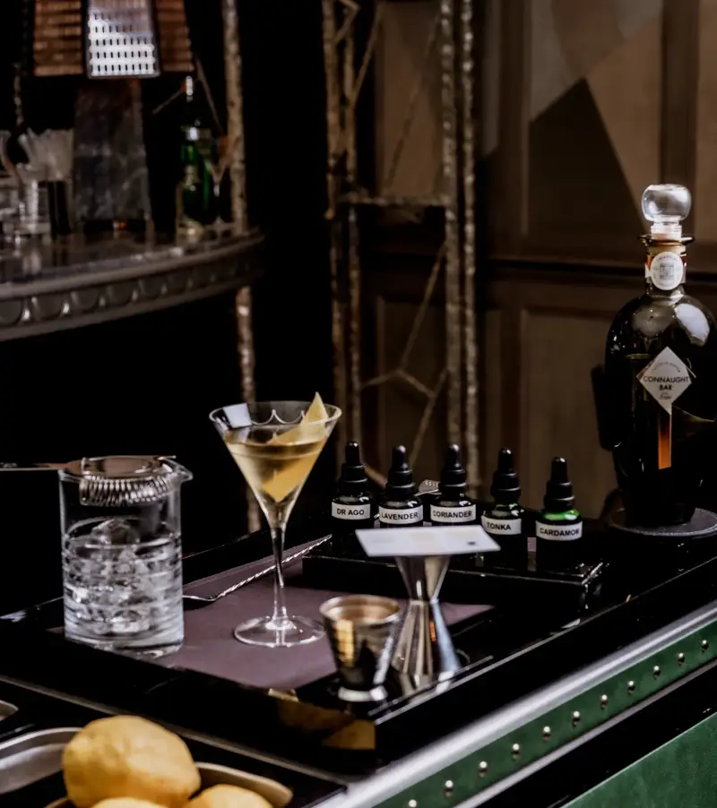 Martini trolley with mixing tools, citrus, bitters bottles, and a prepared martini beside a Connaught Bar gin bottle.
