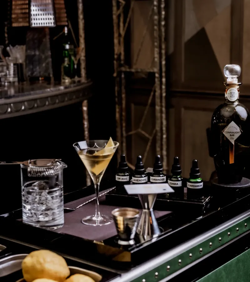 Martini trolley with mixing tools, citrus, bitters bottles, and a prepared martini beside a Connaught Bar gin bottle.