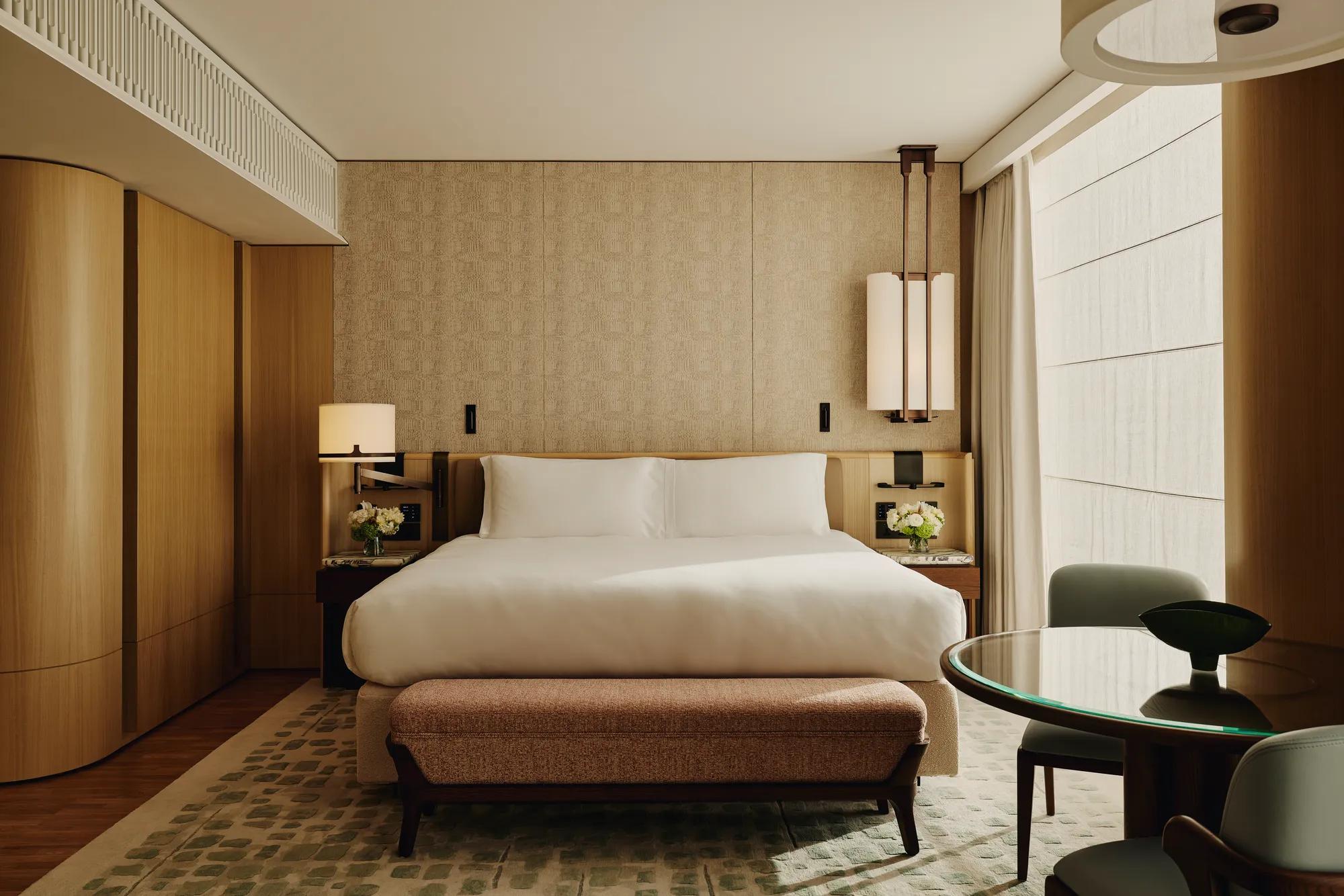 Hotel bedroom with a large bed, patterned wall panels, bedside lamps, a cushioned bench at the foot of the bed, and a round glass-topped table with chairs.