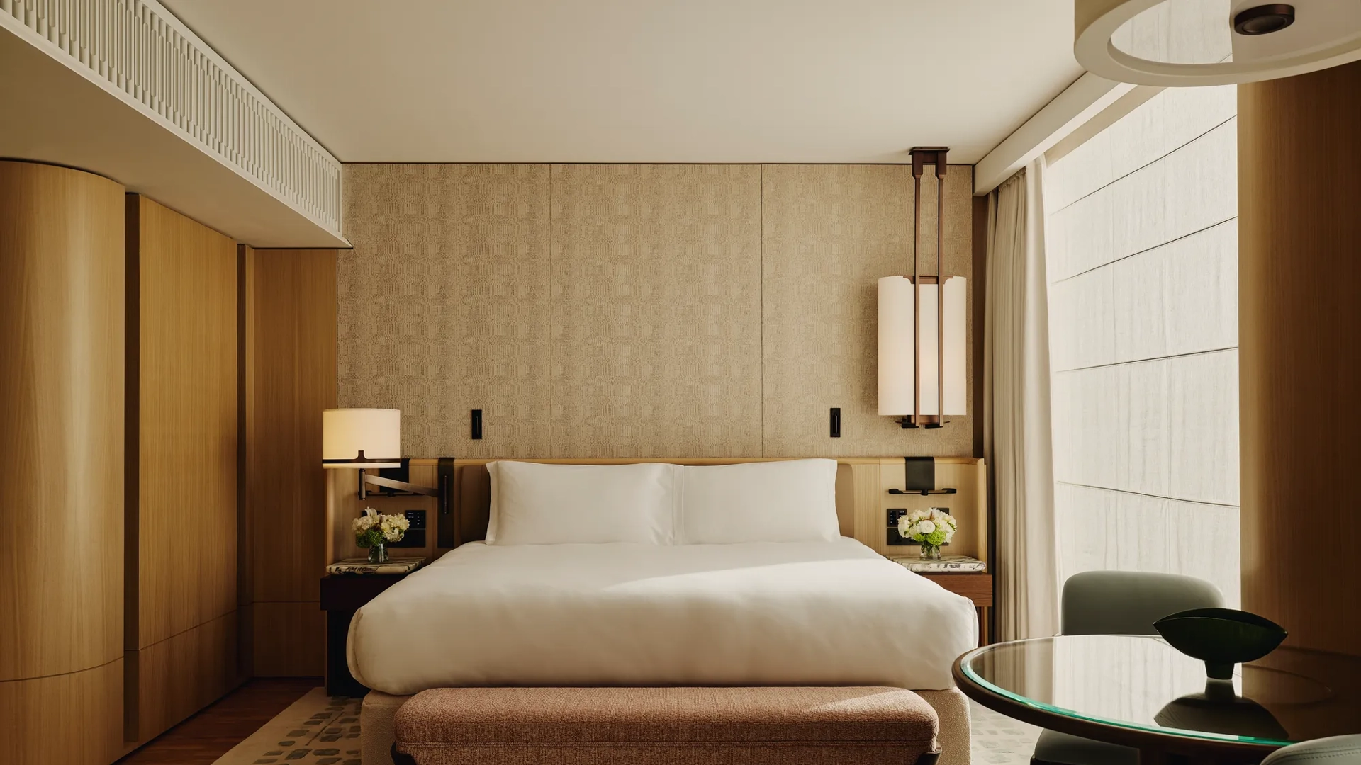 Hotel bedroom with a large bed, patterned wall panels, bedside lamps, a cushioned bench at the foot of the bed, and a round glass-topped table with chairs.