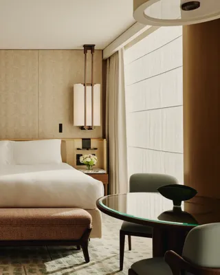 Hotel bedroom with a large bed, patterned wall panels, bedside lamps, a cushioned bench at the foot of the bed, and a round glass-topped table with chairs.