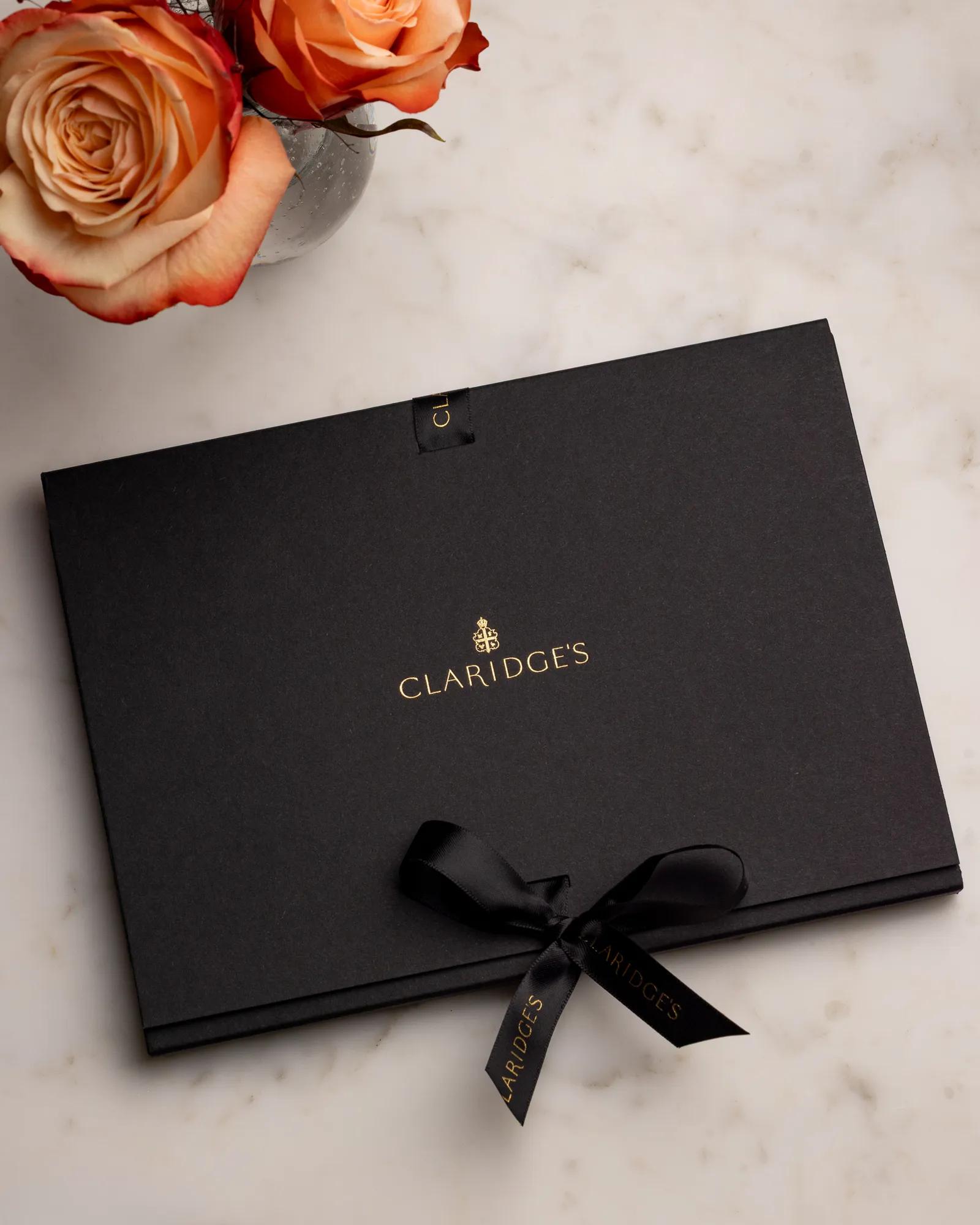 Black Claridge’s gift box with ribbon and gold logo beside a vase of orange roses.
