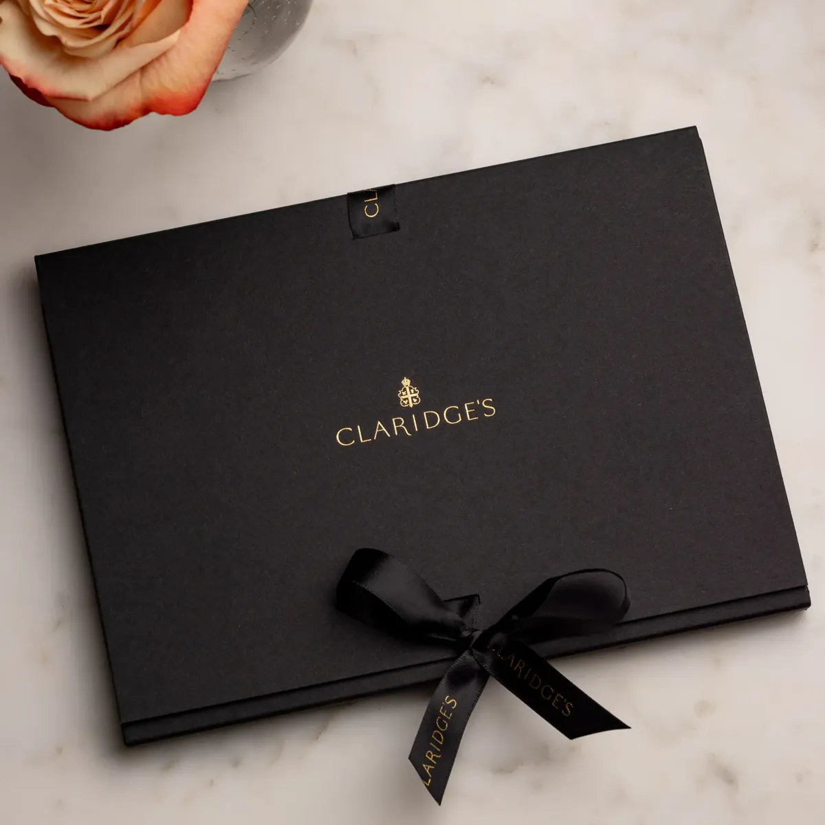 Black Claridge’s gift box with ribbon and gold logo beside a vase of orange roses.