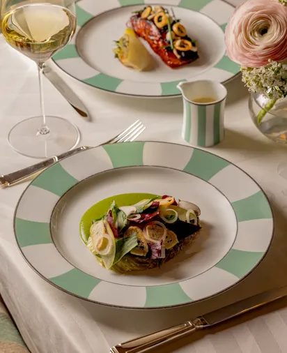 Elegant dining table with two plated dishes on green-and-white patterned china. The foreground plate features fish with green sauce and vegetables, while the background dish shows seared fish with garnish. A glass of white wine and flowers complete the refined setting.