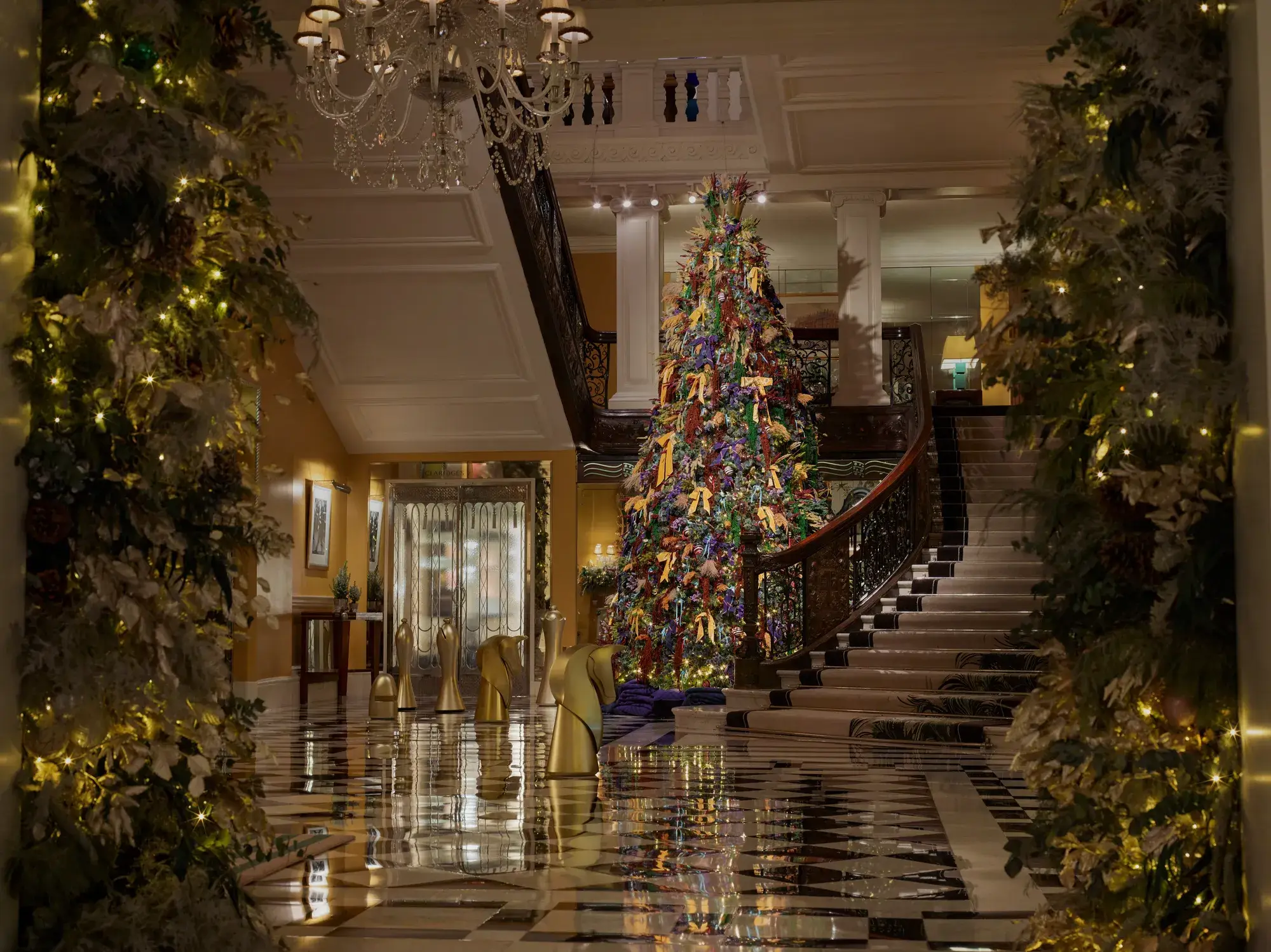 A tall, colourful Christmas tree stands in a grand hotel lobby, framed by sweeping staircases, chandeliers and festive garlands.