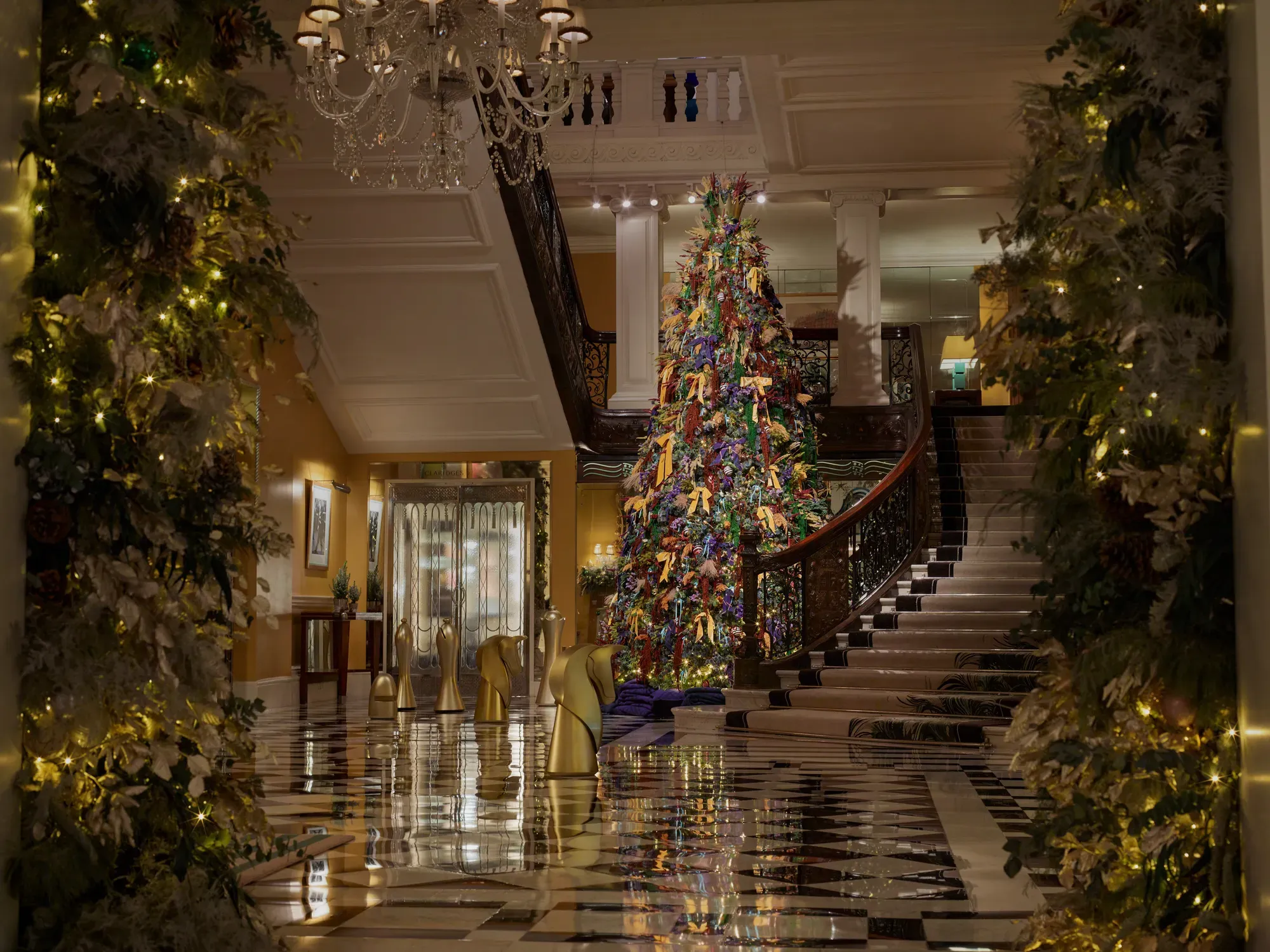 A tall, colourful Christmas tree stands in a grand hotel lobby, framed by sweeping staircases, chandeliers and festive garlands.
