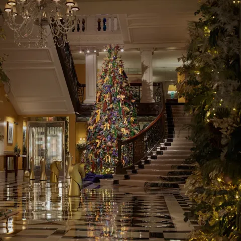 A tall, colourful Christmas tree stands in a grand hotel lobby, framed by sweeping staircases, chandeliers and festive garlands.