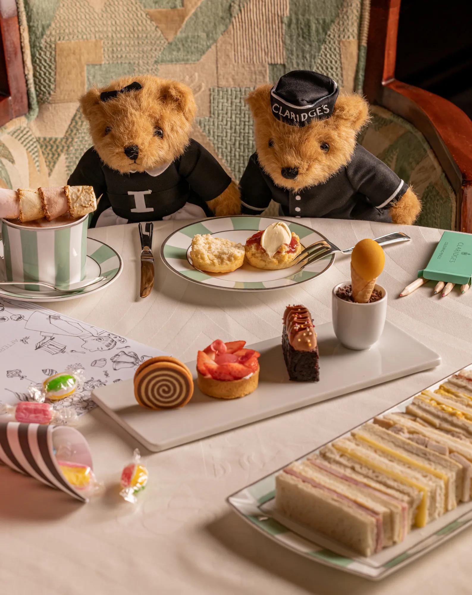 Two teddy bears in Claridge’s outfits seated at a table with afternoon tea pastries, sandwiches and sweets.