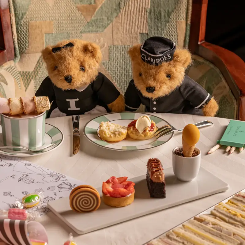 Two teddy bears in Claridge’s outfits seated at a table with afternoon tea pastries, sandwiches and sweets.