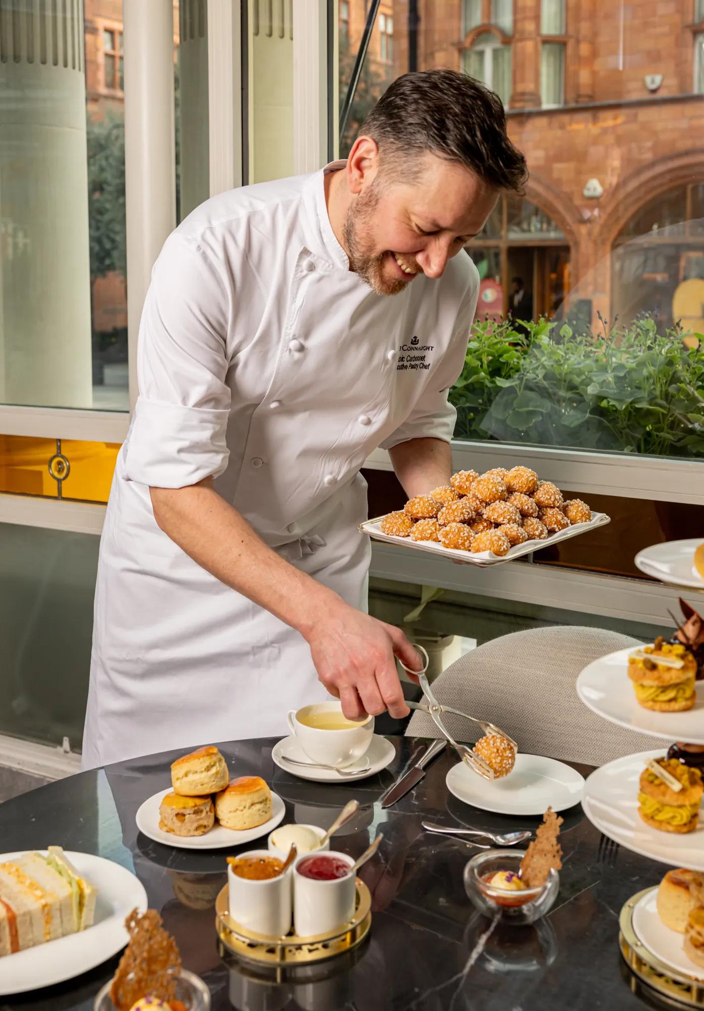 Chef plating pastries and tea at table with scones, sandwiches, and desserts, smiling beside window in bright dining setting.